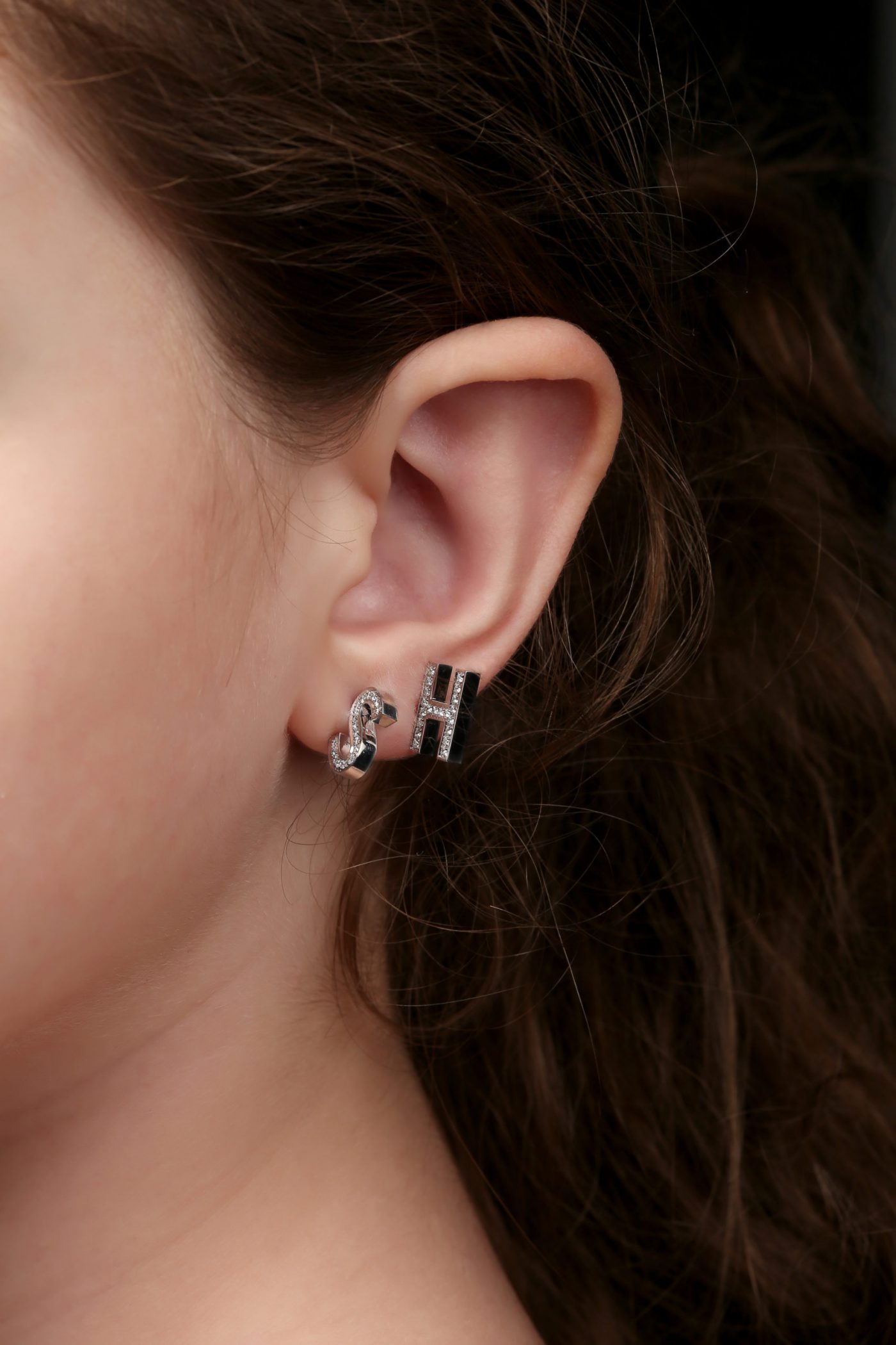 Close-up of silver initial earrings featuring 'S' and 'H' on a model's ear, showcasing modern jewelry design.