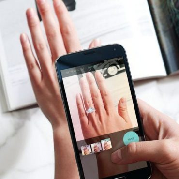 A hand holding a smartphone displaying a virtual try-on feature for a ring on a hand, showcasing jewelry technology.