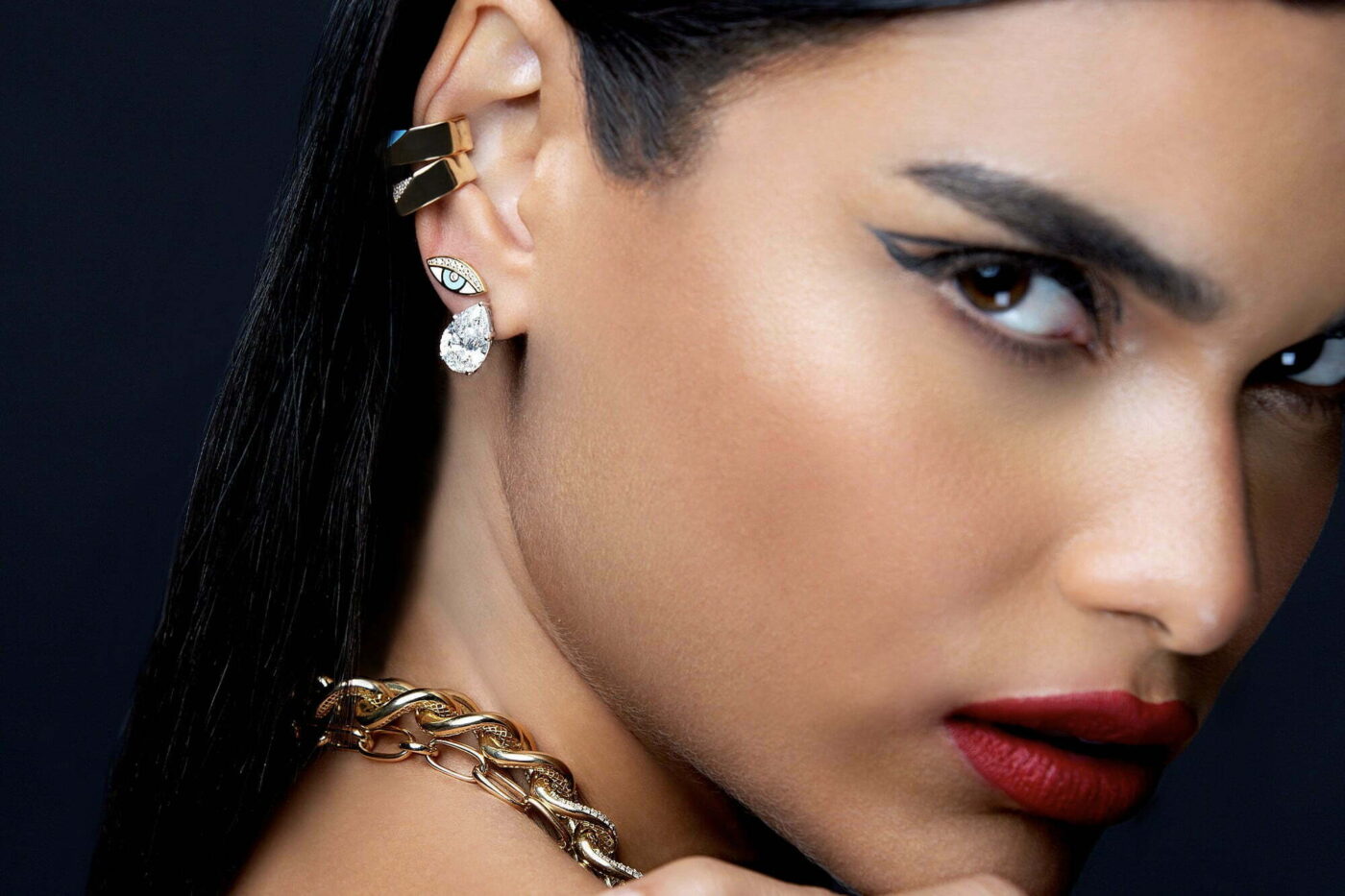 Close-up of a model showcasing high jewelry earrings and a gold chain necklace, featuring a diamond drop and unique design elements.