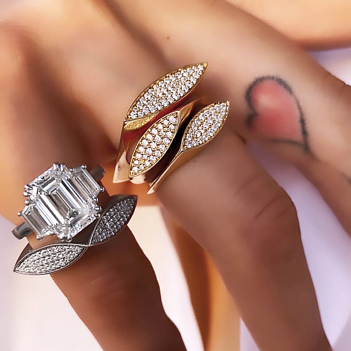 Two rings, one gold with diamonds and one silver with a large emerald-cut stone, displayed on a hand with a tattoo.