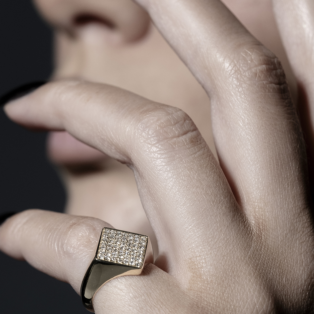 Close-up of a hand wearing a gold ring with a square diamond setting and a person's face partially visible in the background.