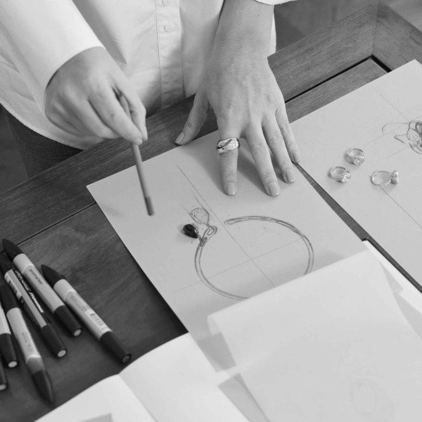 A designer studies jewellery sketches on a worktable, arranging gemstones and rings while refining a hand-drawn piece in black and white tones.